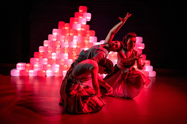 three bharatanatyam dancers striking a sitting pose under red light with a background of white plastic cans.