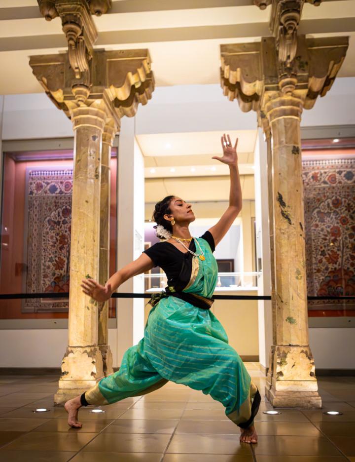 Bharatanatyam artist wearing green saree striking a pose in front of two cream pillars and two persian rugs on the wall behind.
