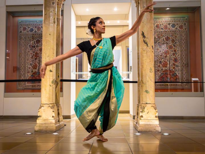 Bharatanatyam artist wearing green saree striking a standing pose in front of two cream pillars and two persian rugs on the wall behind.