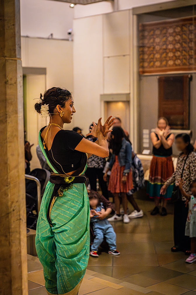 Bharatanatyam artist wearing green saree striking a pose in front of audience including young and old, who are following suit.