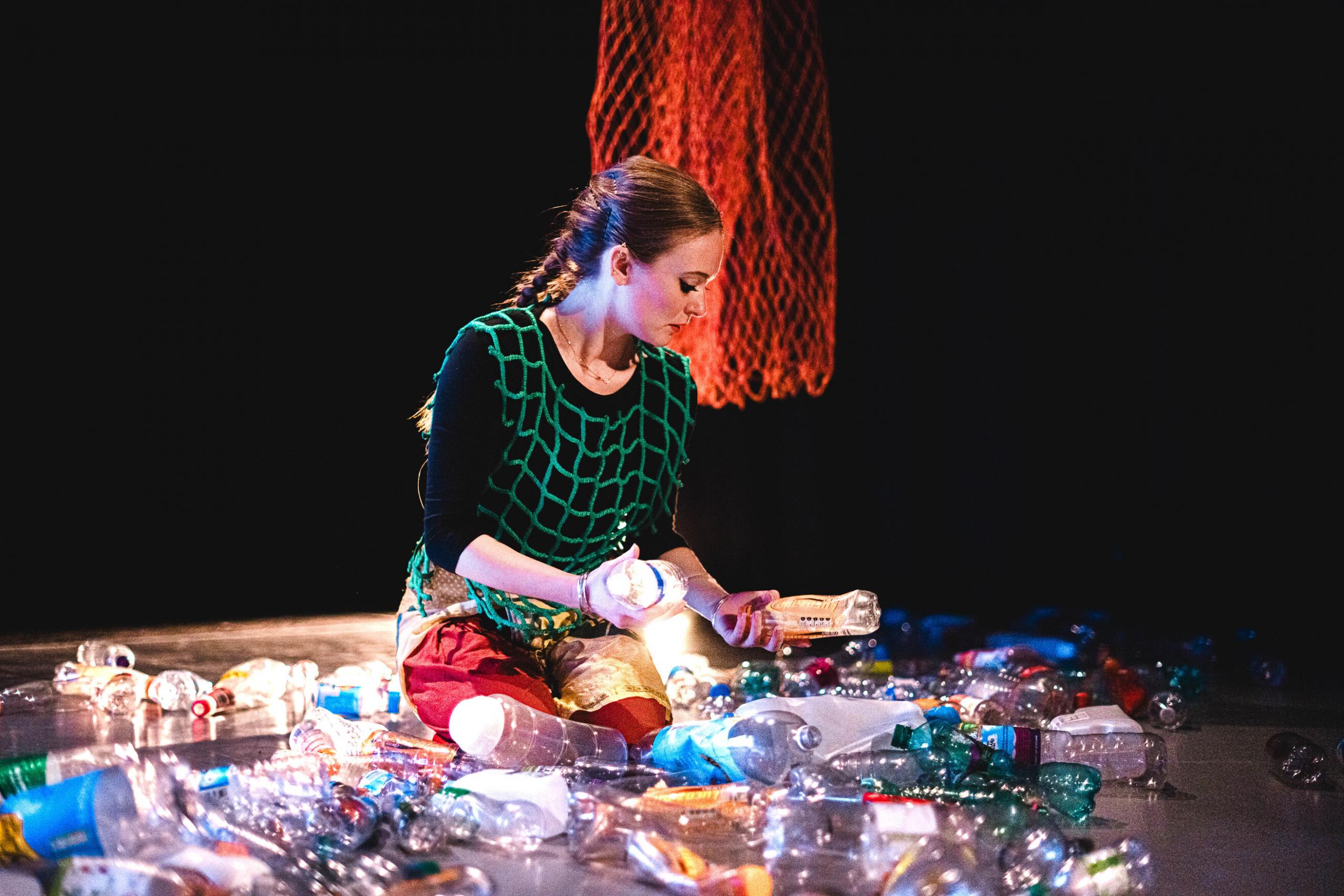 A white female dance artist sitting in the midst of plastic bottles, looking down at them against a black background with an orange fishnet hanging behind her.