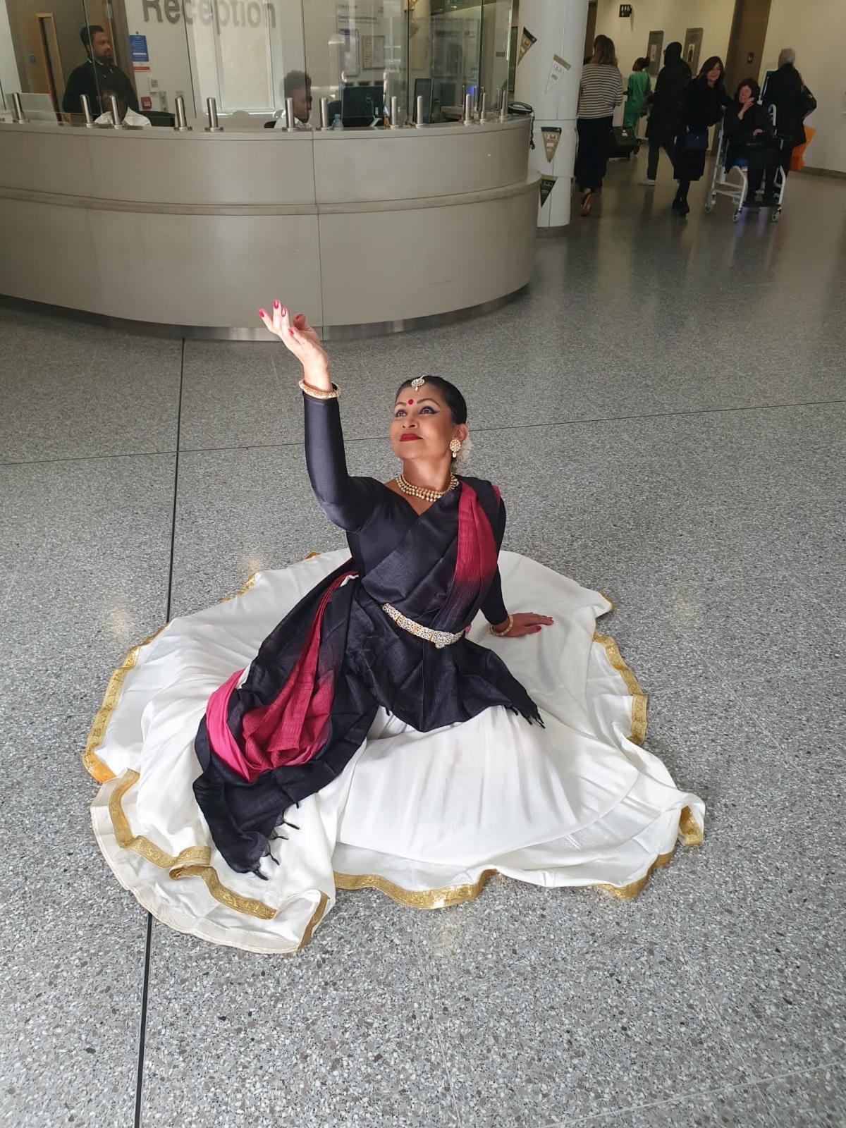 A Kathak dancer sitting on the floor striking a pose. She si dressed in white skirt, black top and a black and red veil, with her hair tied back in a bun.